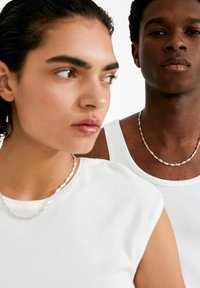 Kaks inimest hõbedaste ketiga kaelakeedega. Naine kannab kehale istuvat valget toppi ja mees valget tanktoppi.