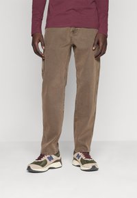 Bruna bomullsshorts med rak klippning, i kombination med sportiga sneakers i flera färger med nät- och mockamaterial. En enkel bakgrund.