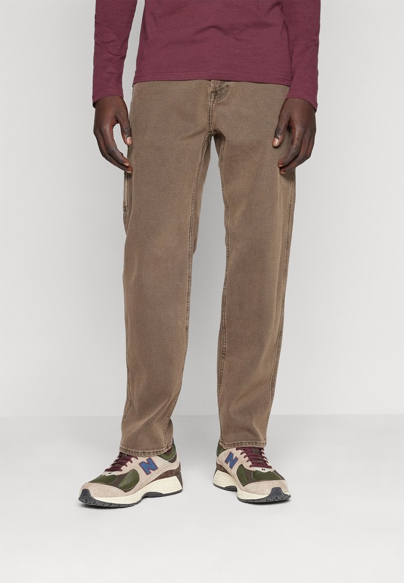 Bruna bomullsshorts med rak klippning, i kombination med sportiga sneakers i flera färger med nät- och mockamaterial. En enkel bakgrund.