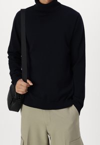 Person i svart polotröja och beige cargo-byxor, som håller i ett svart band kopplat till en liten svart väska över axeln.