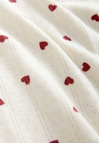 Tissu blanc avec de petits cœurs rouges régulièrement espacés en rangées verticales et des motifs discrets en broderie ajourée entre les cœurs.