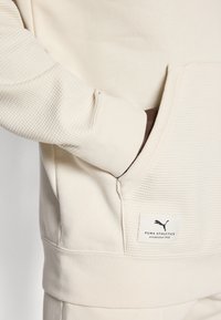 Cremefarvet sweatshirt med teksturerede ærmer, lomme foran og et lille PUMA-logo ved bunden. Blødt, tykt stof.