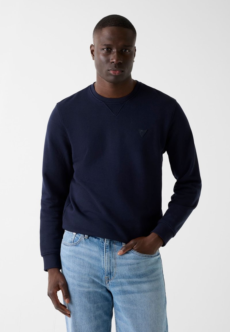 Sweat-shirt bleu marine avec col rond, poignets côtelés et logo triangulaire minimaliste sur la poitrine. Associé à un jean en denim bleu clair.