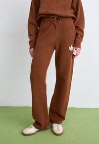 Braune, weiche, gestrickte Jogginghosen mit einem Tunnelzug und besticktem Logo-Detail. Kombiniert mit beigen und braunen Sportschuhen.
