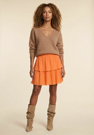 Frau mit lockigem Haar, die einen braunen V-Ausschnittpullover, einen orangefarbenen gestuften Rock und lässige beige Stiefel vor einem einfarbigen Hintergrund trägt.