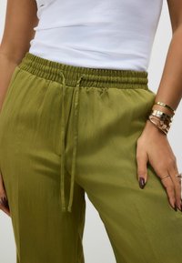 Olivgrüne Hosen mit elastischem Bund und Kordelzug. Weicher Stoff mit lockerer Passform und seitlichen Taschen. Mehrere metallische Armbänder sind sichtbar.