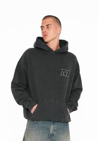 Tmavě šedá mikina s kapucí vyrobená z měkkého materiálu, s přední kapsou a vyšitým detailem "M". Má uvolněný střih a velkou kapuci.