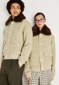 Beige plüssdzsekik barna szőrme gallérral, nagy gombokkal és elülső zsebekkel. Az egyik modell csíkos nadrágban, a másik kockásban van.