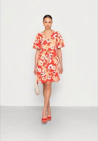 Vestido floral vermelho com mangas curtas e volumosas, cintura ajustada e saia até ao joelho. Combinado com sapatos de salto vermelho e uma bolsa de mão bege claro.