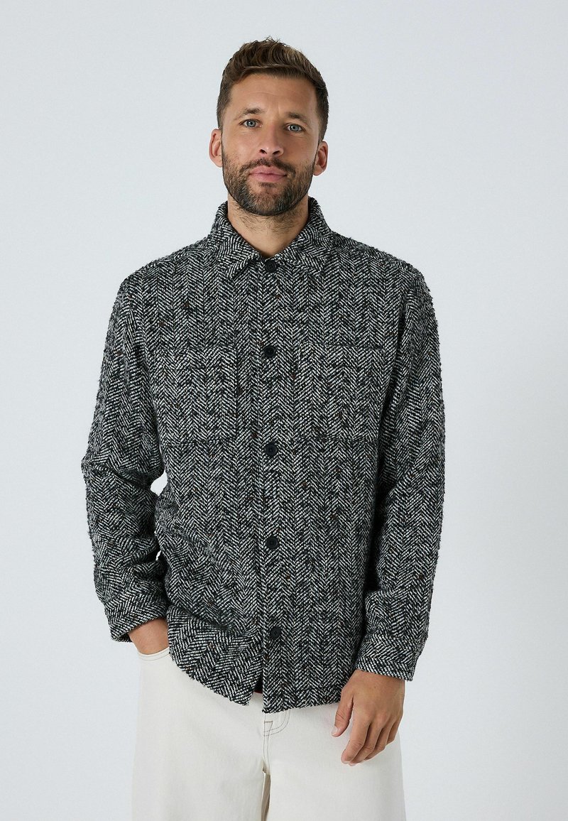 Košeľa s herringbone vzorom v čierno-bielej farbe, vyrobená z textúrovanej látky, so golierom a prednými vreckami.