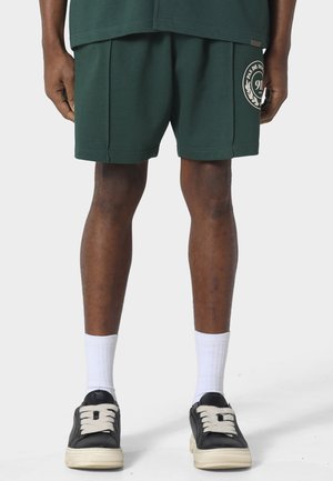 Osoba ubrana w ciemnozielone spodenki z logo na lewej nodze, białe skarpetki do kostek oraz czarne trampki z białą podeszwą i sznurowadłami.