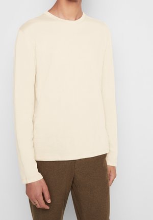 Mężczyzna ubrany w kremową, długorękawową koszulkę z okrągłym dekoltem oraz brązowe spodnie o fakturze, stojący na tle jednolitego, jasnego tła.