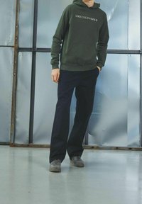 Zielona bluza z kapturem z napisem "UNDISCOVERED" na białym tle, połączona z ciemnymi spodniami i szarymi butami, na metalicznym tle.