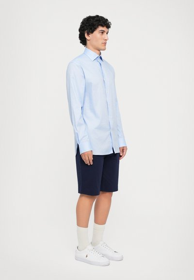 Ljusblå långärmad skjorta med knappar och spetsig krage, kombinerad med marinblå shorts. Modellen bär vita sneakers och krämfärgade strumpor.