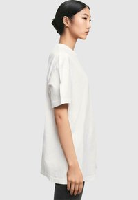 Weißes Baumwoll-T-Shirt, kurze Ärmel, Rundhalsausschnitt, lockere Passform, gestickte Nähte, getragen mit dunklen Hosen, vor einem neutralen Hintergrund präsentiert.