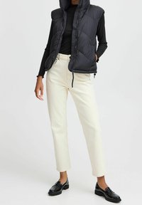 Czarna kamizelka puchowa, zapinana na zamek, z wysokim kołnierzem, noszona na dopasowanej czarnej bluzce. Jasnobeżowe proste spodnie i czarne mokasyny dopełniają outfit.