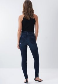 Jeans ajustados de color azul oscuro con una textura suave, cintura alta y costura sutil en los bolsillos traseros, combinados con una blusa negra sin mangas.