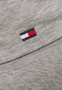 Grauer Stoff mit ribbeliger Textur zeigt ein kleines gesticktes Logo in Marineblau, Rot und weißen Streifen in der Nähe der Naht.