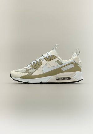 Zapatilla Nike Air Max en beige y blanco con el símbolo swoosh blanco, diseño con cordones, amortiguación visible en la suela y tiradores en la lengüeta y el talón.