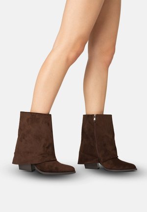 Bruine suède laarzen tot halverwege de kuit met zijrits en lage blokhakken, gedragen op blote benen tegen een effen lichte achtergrond.
