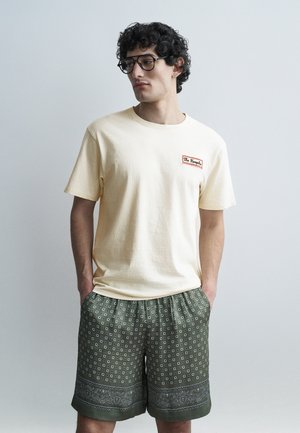 Jeune homme aux cheveux bouclés et lunettes portant un t-shirt crème et un short vert olive à motifs, les mains dans les poches, regardant sur le côté.