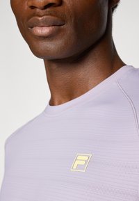 Camiseta atlética de manga corta color púrpura claro con una superficie texturizada y un pequeño logo dorado en el pecho. Presenta un cuello redondo y un diseño sin costuras.