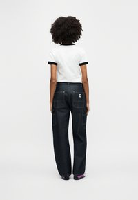 Άτομο με σγουρά μαλλιά που φοράει λευκό μπλουζάκι με μαύρο περίγραμμα και σκούρο μπλε τζιν παντελόνι φαρδύ με πολλές τσέπες, στέκεται με την πλάτη γυρισμένη.