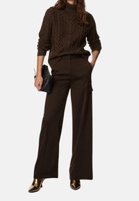 Brązowy sweter o splocie warkoczowym z wysokim kołnierzem, w połączeniu z szerokimi brązowymi spodniami. Dodatki to czarna torebka i brązowe botki za kostkę.