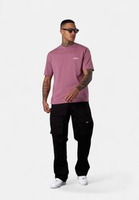 Mann mit Tattoos und Sonnenbrille, der ein mauvefarbenes T-Shirt, schwarze Cargo-Hosen und weiße Sneakers trägt und vor einem schlichten weißen Hintergrund steht.