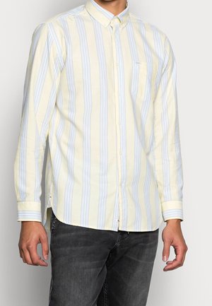 Man draagt een geel-wit gestreept overhemd met lange mouwen en zwarte jeans, staand tegen een witte achtergrond.