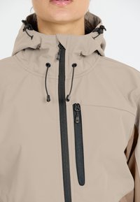 Beige jacket with a high collar, adjustable drawstrings, and black zippers. Features a side zip pocket and a smooth, weather-resistant fabric.