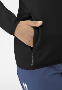 Black fabric pullover with a zippered hand pocket, featuring a textured surface. Blue trousers with a white logo at the hem.