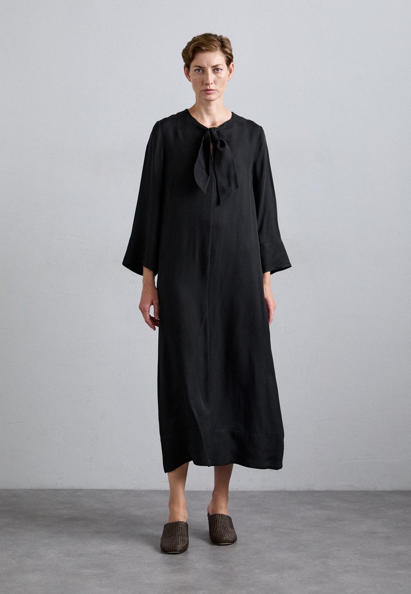 Black long-sleeve dress made of smooth fabric, featuring a bow tie at the neck and a straight silhouette. Worn with brown textured slip-on shoes.