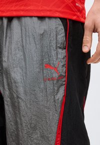 Κοντινό πλάνο ατόμου που φοράει γκρι και μαύρα σορτς Puma με κόκκινο περίγραμμα και λογότυπο Slam Jam, καθώς και κόκκινο αθλητικό πουκάμισο.