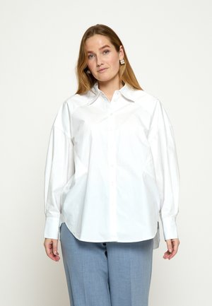 Žena s dlhými vlasmi, oblečená v nadrozmernej bielej košeli a svetlosivých nohaviciach, stojaca pred jednoduchým svetlým pozadím.