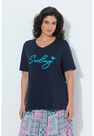 Kobieta o kręconych, ciemnych włosach nosi granatowy T-shirt z napisem „Smiling” w kolorze morskim oraz spódnicę w geometryczne wzory.