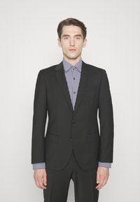 Completo nero su misura con due bottoni, rever a punta e tasche laterali, abbinato a una camicia a fantasia blu e bianca con dettagli a bottoni.