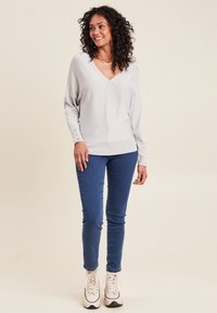 Helles graues V-Ausschnitt-Pullover mit langen Ärmeln und Knopfakzenten an den Bündchen, kombiniert mit schmal geschnittenen blauen Jeans und beigen Sneakers.