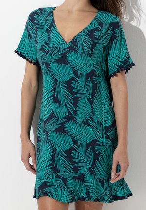 Donna che indossa un vestito corto blu navy con stampa di foglie di palma verde brillante, rifinitura con pon pon sulle maniche e orlo arricciato, in piedi contro un muro bianco.