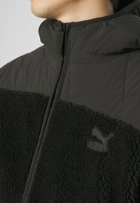 Veste noire avec une partie supérieure matelassée et une partie inférieure douce. Fermeture éclair, col montant et accent logo subtil sur la texture.
