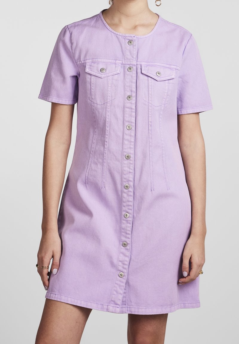 Robe en denim lilas avec manches courtes, deux poches poitrine et fermeture à boutons sur le devant. Présente des détails de surpiqûres et une silhouette ajustée.