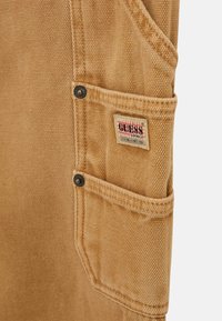 Beige Stofftasche mit grober Textur, versehen mit einem Etikett mit rotem und schwarzem Text. Enthält schwarze Metallknöpfe und mehrere Taschen.
