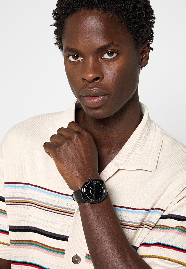 Reloj de pulsera negro con esfera redonda y correa texturizada; el reloj tiene detalles plateados y se lleva en una muñeca frente a una camisa de rayas.