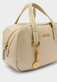 Beige Lederhandtasche mit strukturiertem Design, oberen Griffen, goldfarbenen Beschlägen und einem dekorativen Charm. Reißverschlussverschluss mit subtiler Textur.