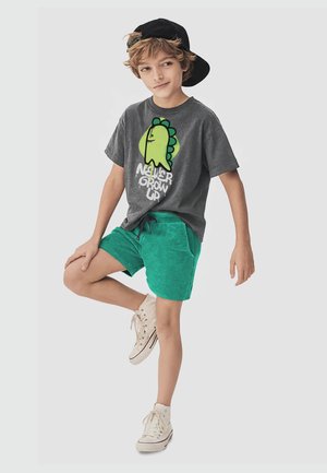 Boy wearing a backward black cap, gray "Never Grow Up" t-shirt with green dinosaur, green shorts, and white sneakers, balancing on one leg.