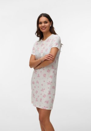 Γυναίκα που φοράει λευκό φόρεμα με κοντά μανίκια και ροζ λουλουδάτα μοτίβα, στέκεται με σταυρωμένα τα χέρια και χαμογελάει μπροστά σε απλό λευκό φόντο.