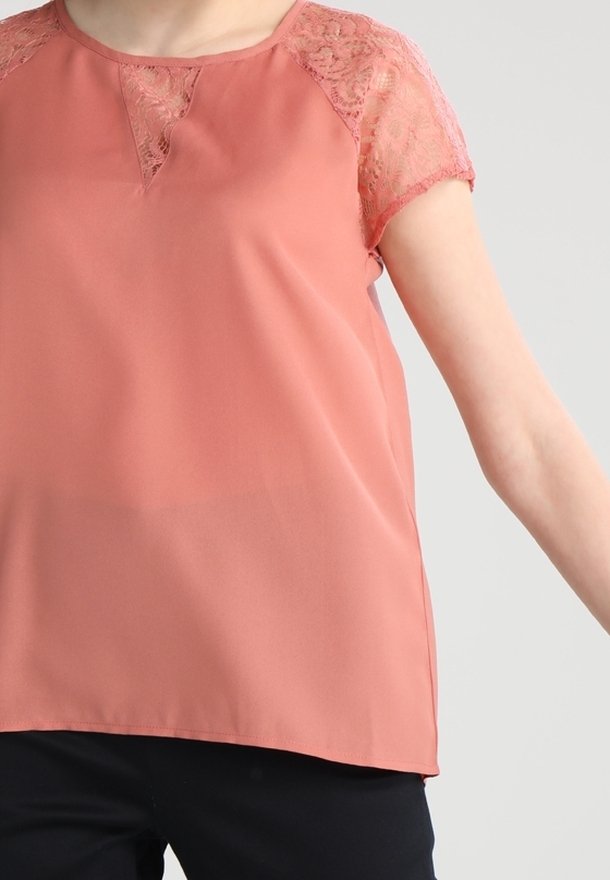 Femme portant un chemisier rose saumon avec des manches en dentelle et un col, associé à un pantalon noir, se tenant avec un bras tendu.