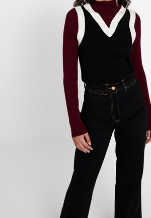 Femme portant un haut noir sans manches à col en V avec des accents blancs sur un col roulé côtelé bordeaux et un pantalon noir taille haute.