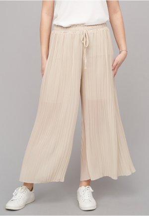 Mujer con pantalones beige de pierna ancha y plisados con cintura de cordón y zapatillas blancas sobre un fondo gris.