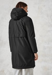 Zwarte jas van gemiddelde lengte met een capuchon, met een gladde textuur en een ritszak aan de zijkant, ontworpen met een ontspannen pasvorm en een iets getailleerde taille.